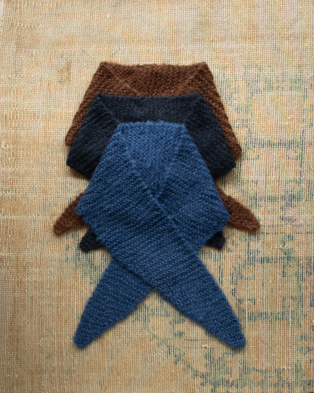Three knitted scarves in brown, blue, and navy on a textured beige surface.